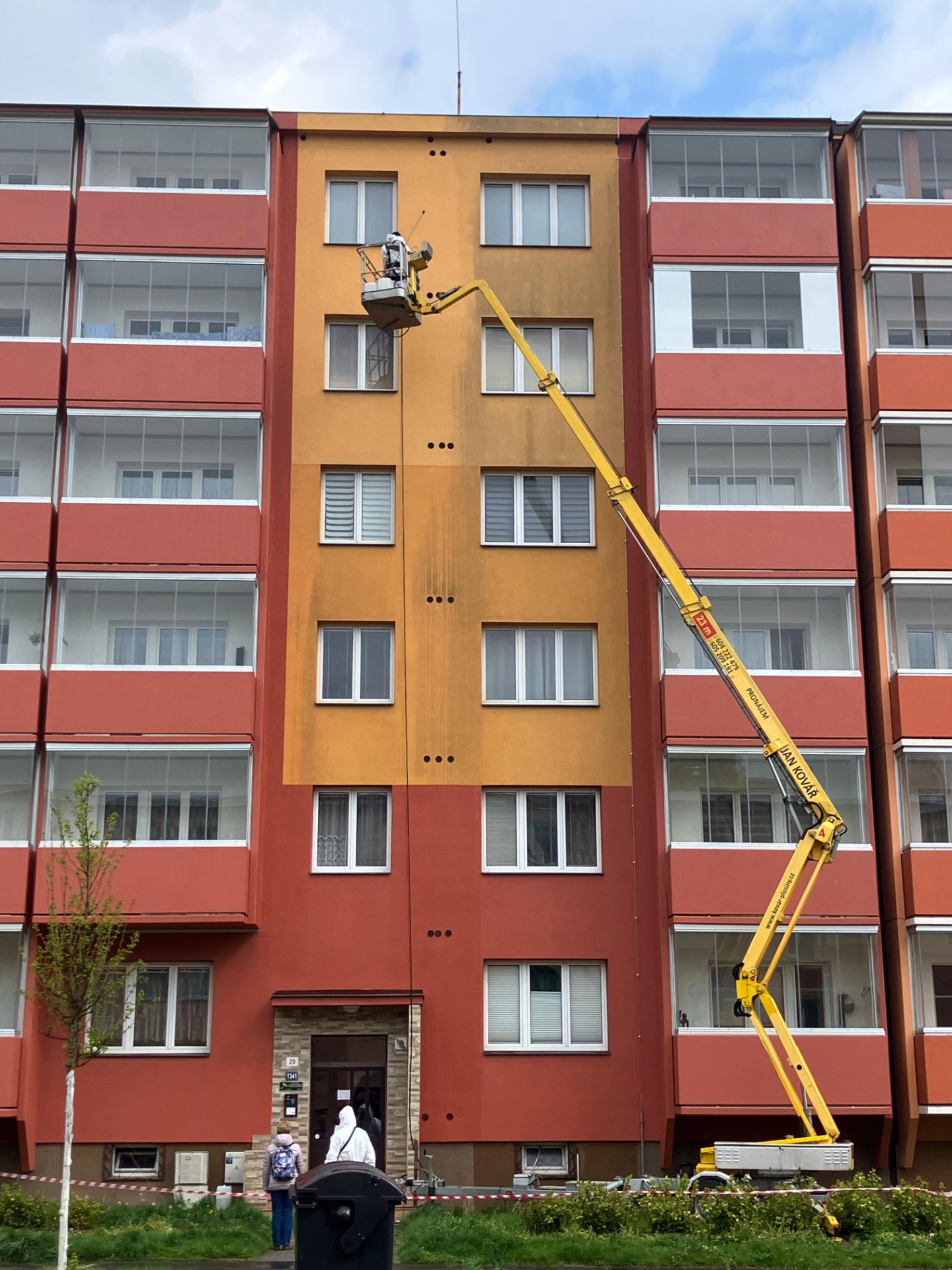 Žlutý teleskopický jeřáb s košem pracuje u fasády bytového domu mezi okny. Výškové práce s pracovní plošinou nebo z lana. 