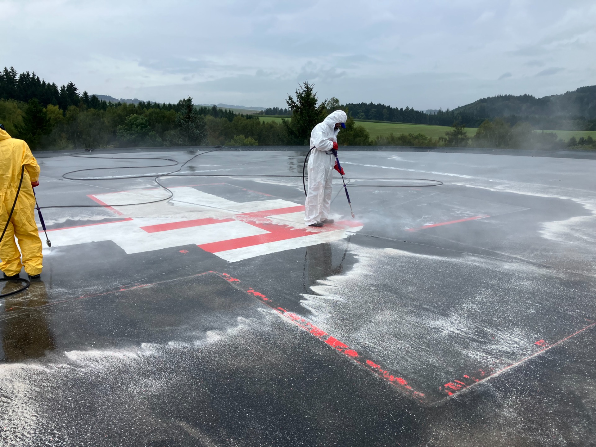 Pracovník v ochranném obleku stříká dezinfekci na mokrou asfaltovou plochu s červenými značkami. Tryskání heliport pro odstranění barvy z asfaltu