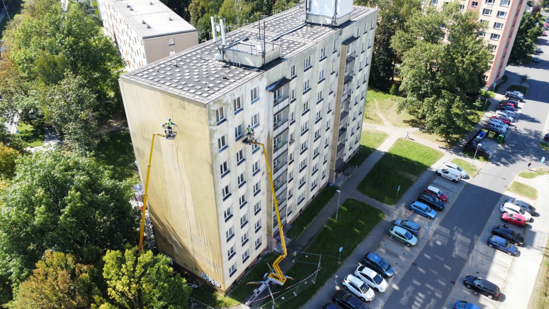 Výškové práce z plošiny, kdy pracovník pracuje uprostřed fasády bytového domu, čistí špinavou fasádu vysokotlakým čištěním ve výšce