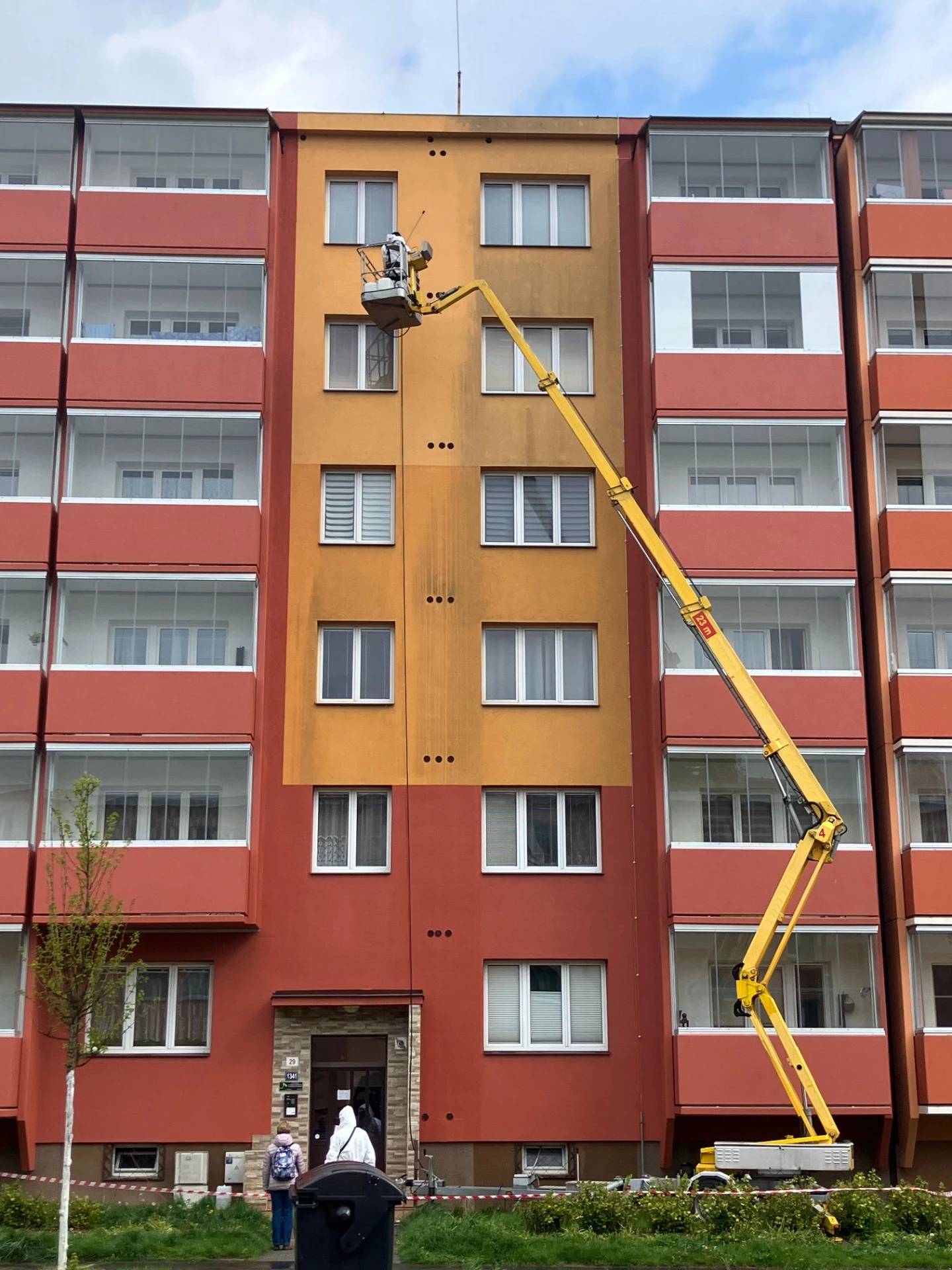 Žlutý teleskopický jeřáb s košem pracuje u fasády bytového domu mezi okny. Výškové práce s pracovní plošinou nebo z lana. 
